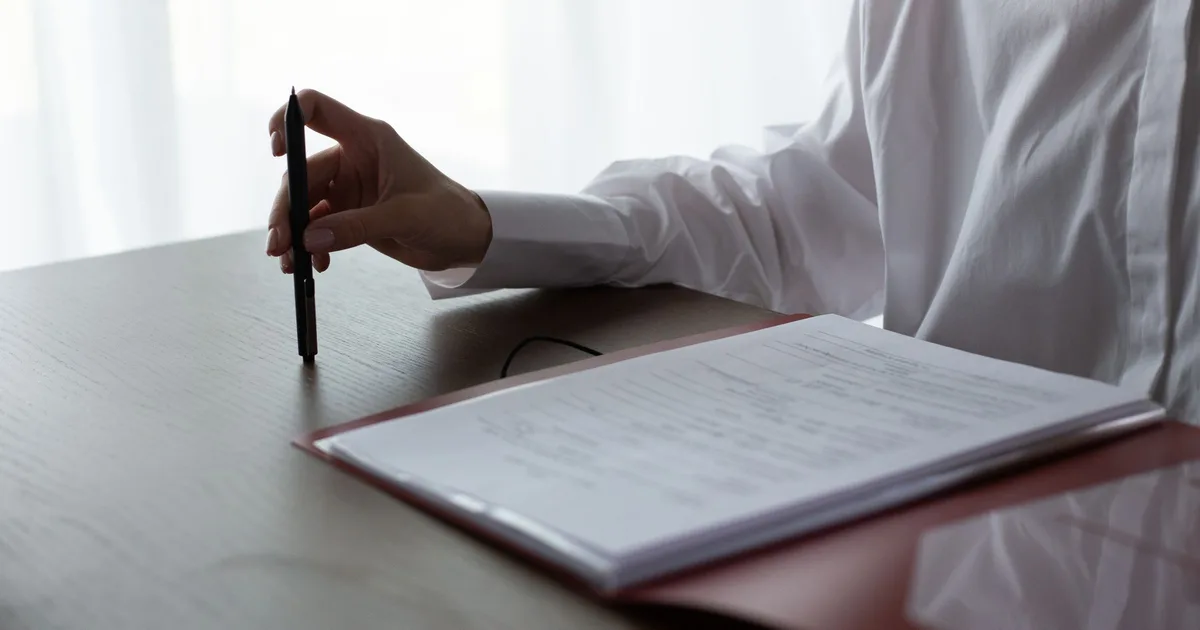 A person in a white shirt holds a pen beside a folder of documents on a wooden desk